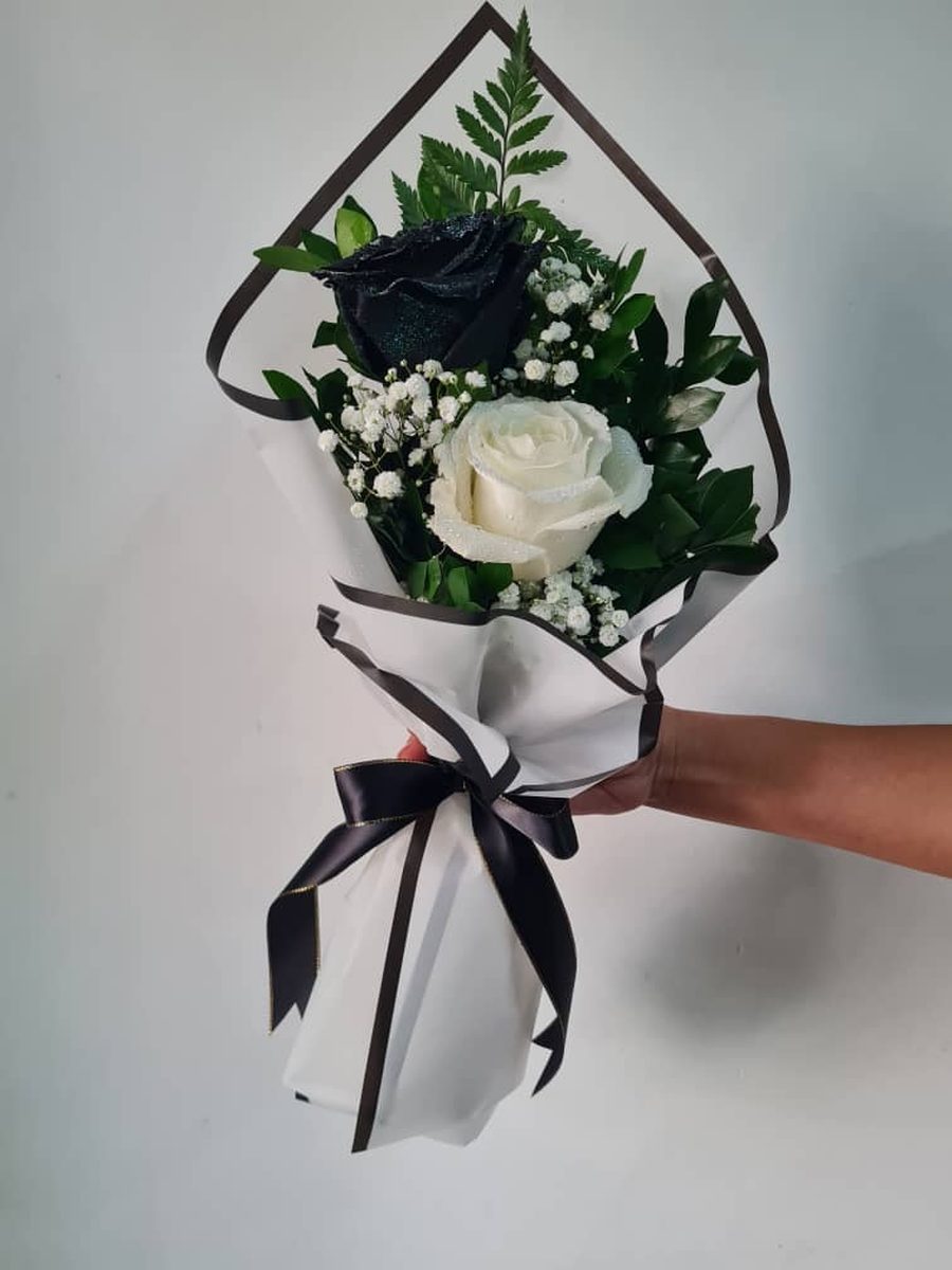 Dúo de rosas: una rosa negra y una blanca con helecho, envueltas en blanco y negro